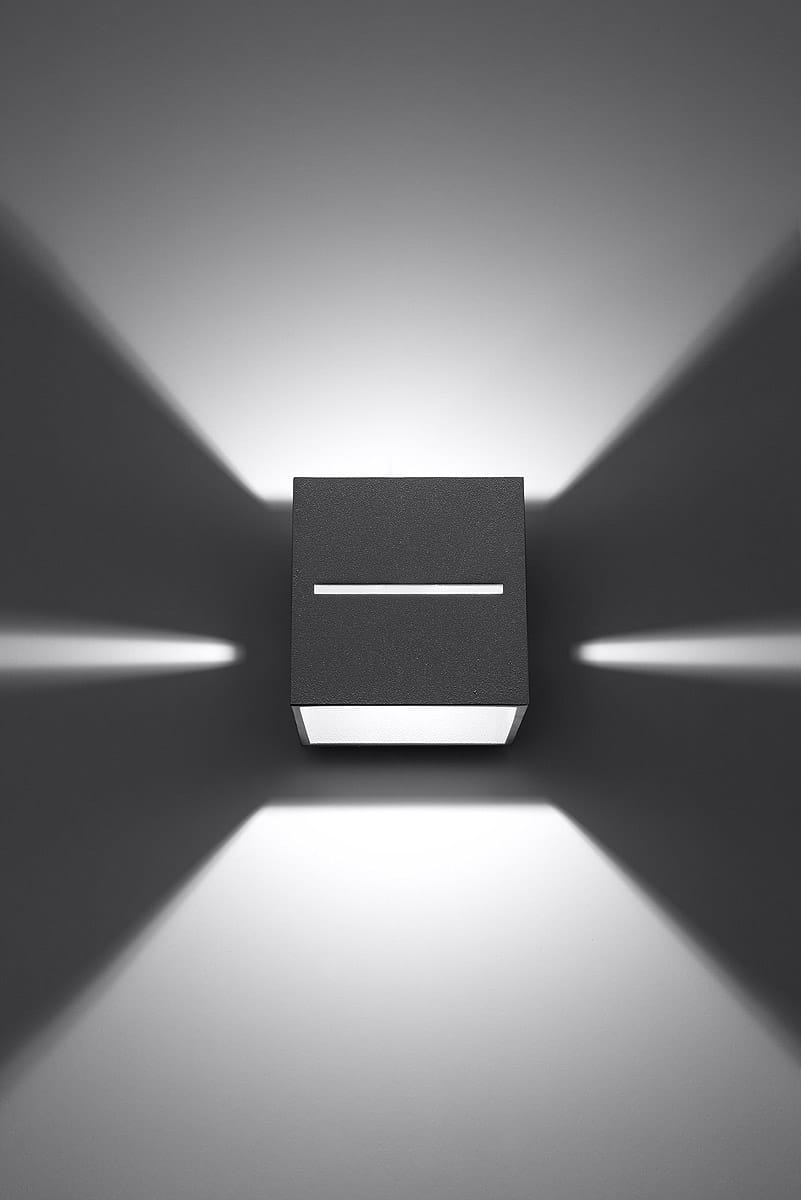 Kinkiet Lobo 1-punktowy w kolorze szarym, o prostokątnej formie. Zamontowany na ścianie, emitujący światło w górę i w dół, tworząc ciekawe cienie. Obudowa matowa, z białym kloszem na dole, z subtelną, poziomą szczeliną na froncie.