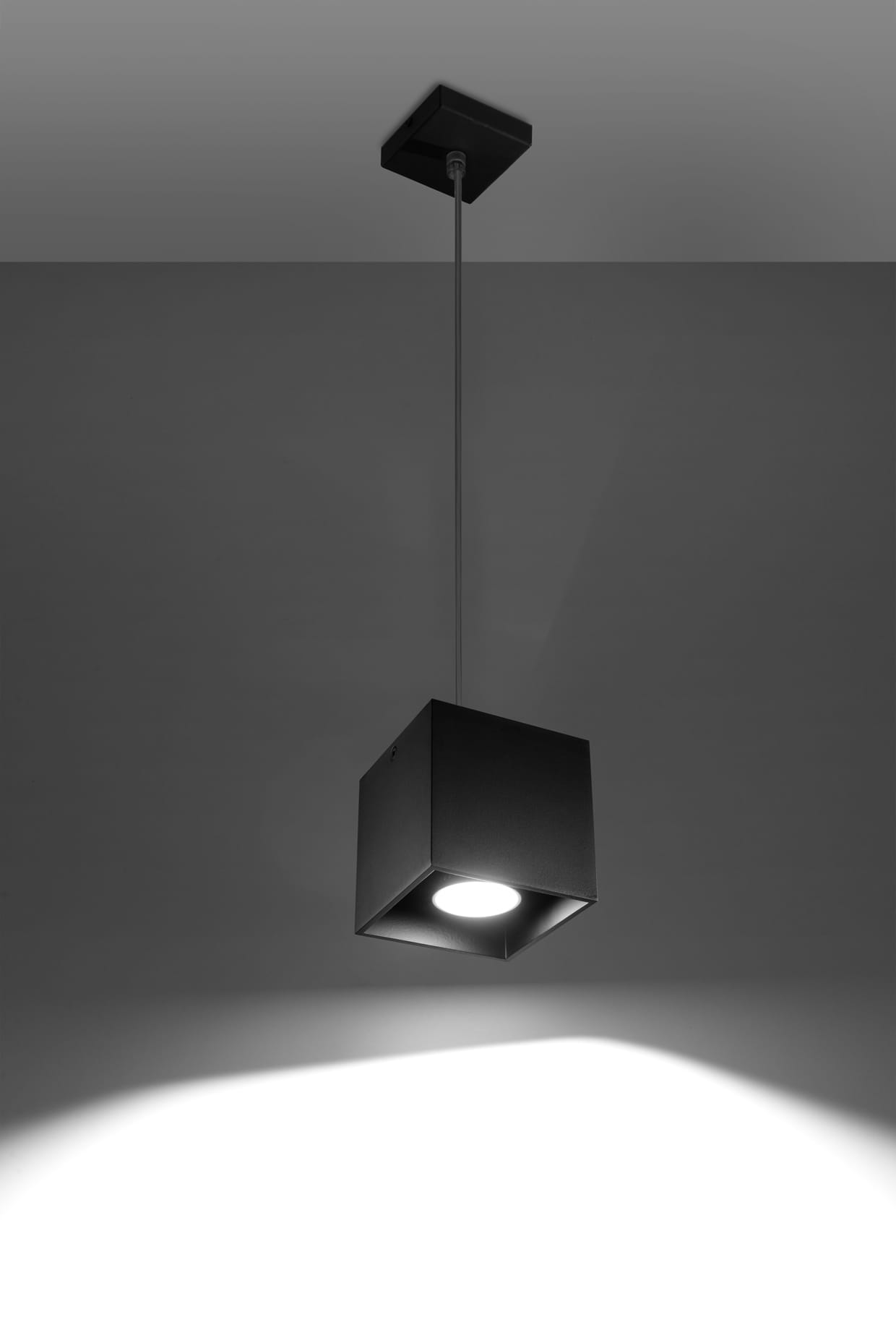 Lampa wisząca Quad 1-punktowa w kolorze czarnym, w formie sześcianu, zawieszona na cienkim przewodzie. Emituje światło z centralnego źródła, na tle szarej ściany, tworzy cień w kształcie prostokąta.