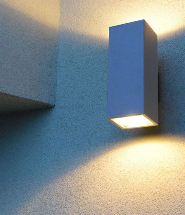 Kinkiet elewacyjny Adela 2-punktowy wykonany z aluminium, w kształcie prostokątnym. Montowany na ścianie, emituje światło w górę i w dół. Kolor szary, z widoczną, szklana osłoną u dołu. Tło w odcieniu niebieskim.