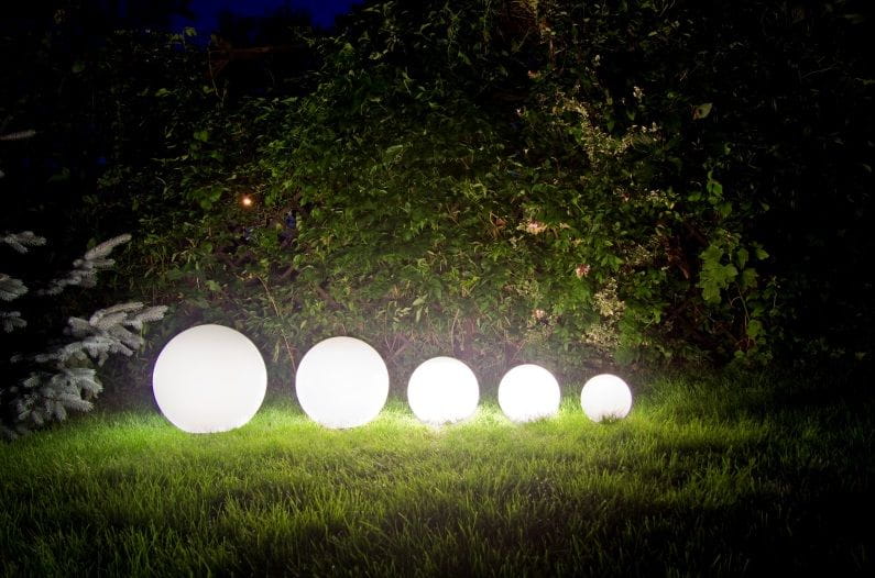 Lampa stojąca ogrodowa Alekule G 200 Z w formie białych, okrągłych kul różnej wielkości, ustawionych na zielonej trawie, z tłem z roślinności. Oświetlenie daje efekt miękkiego światła w ciemności.