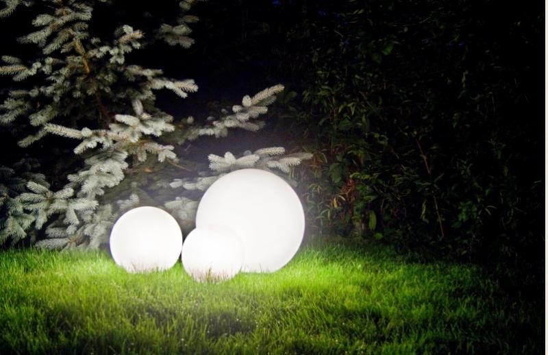 Lampa stojąca ogrodowa Alekule G 250 Z w formie trzech kul o białej, matowej powierzchni, różnej wielkości, umieszczona na trawie w ciemnym otoczeniu. Tło stanowi zieleń krzewów, co podkreśla jej dekoracyjny charakter.