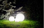 Lampa stojąca ogrodowa Alekule G 250 Z w formie trzech kul o białej, matowej powierzchni, różnej wielkości, umieszczona na trawie w ciemnym otoczeniu. Tło stanowi zieleń krzewów, co podkreśla jej dekoracyjny charakter.