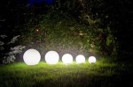 Lampa stojąca ogrodowa Alekule G 500 Z, z okrągłym, białym kloszem, umieszczona na trawie. W tle widoczne zielone krzewy. Produkt o różnej wielkości kulach, emitujący miękkie światło, ustawiony na prostym tle.