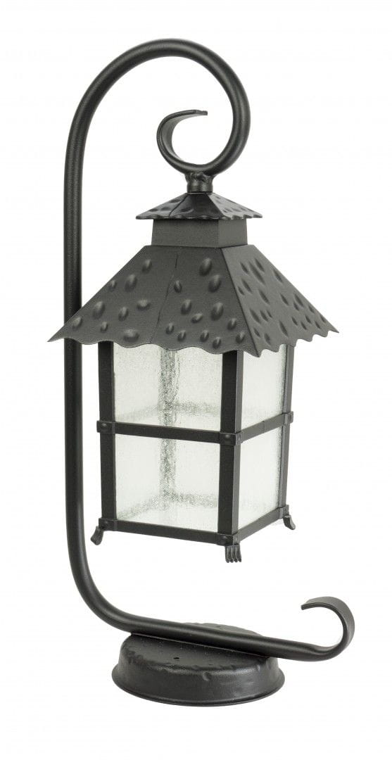 Czarna lampa stojąca ogrodowa Cadiz z metalowym stelażem i szklanym kloszem. Posiada dach w kształcie piramidy z wypukłymi detalami. Ujęcie frontalne, lampa umieszczona na okrągłej podstawie, z giętym uchwytem.