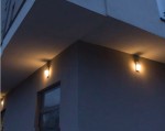 Kinkiet elewacyjny City z aluminium, 1-punktowy, zamontowany na ścianie w rogu budynku. Oświetlenie emituje ciepłe światło, widoczne na szarej elewacji. Produkt ma prostokątny kształt z dekoracyjnym wzorem na obudowie.
