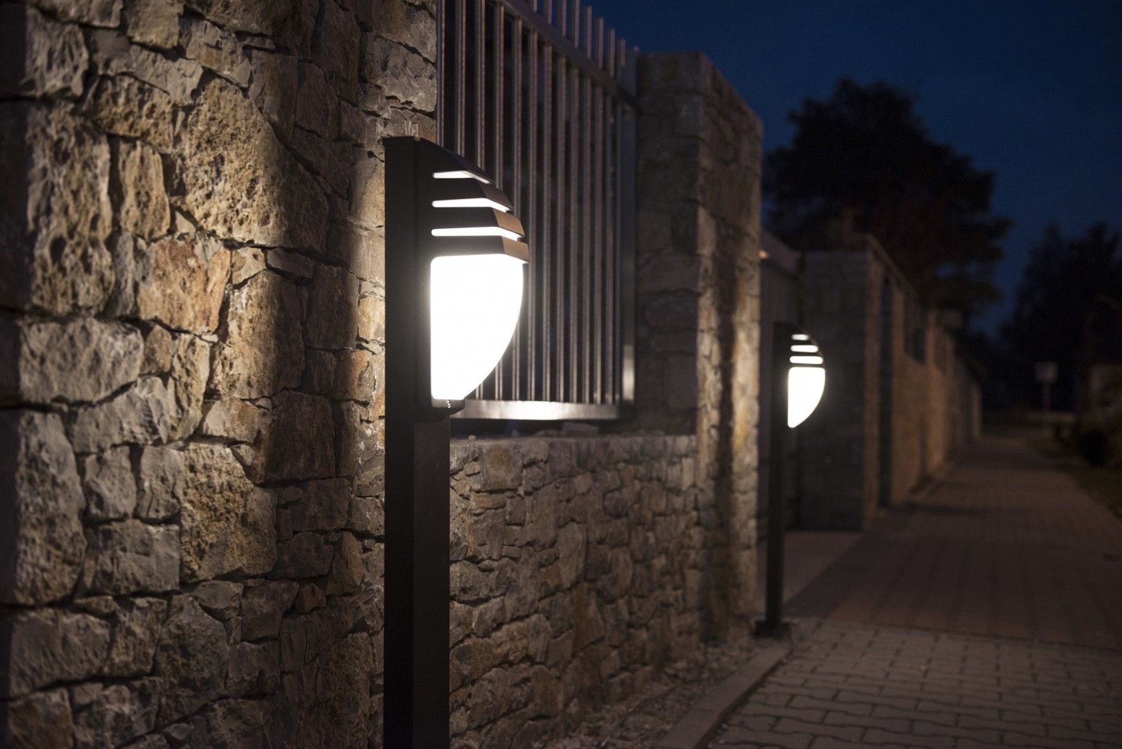 Czarna lampa stojąca ogrodowa City 1-punktowa, o okrągłej, białej kloszowej głowicy z dekoracyjnymi szczelinami. Ujęcie z boku, w nocy, przy kamiennej ścianie, podkreślająca stylowe oświetlenie przestrzeni zewnętrznej.