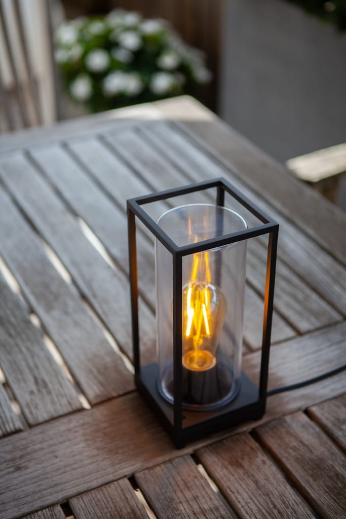 Czarna lampka stojąca zewnętrzna Flair z obudową z metalu i przezroczystym kloszem. Widoczna żarówka Edison, emitująca ciepłe światło. Ujęcie z góry na drewnianym stole, z kwiatami w tle.