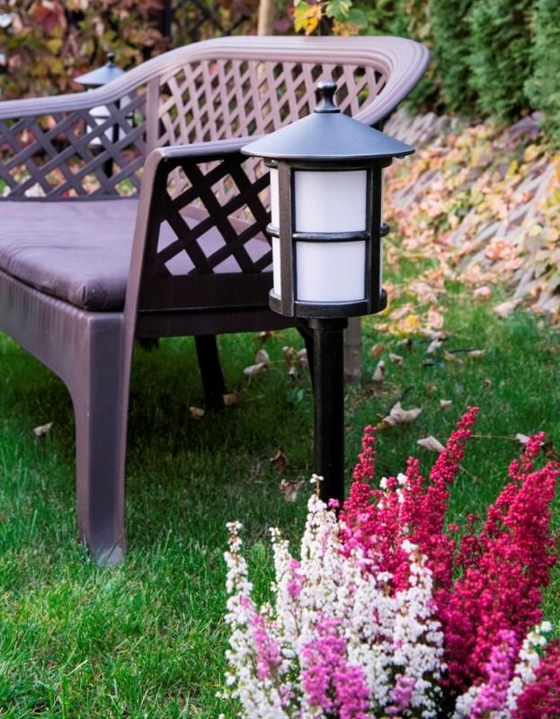 Lampa stojąca ogrodowa Cordoba H71 II, czarna, z białym kloszem, umieszczona obok brązowej kanapy na trawie. W tle widoczne kolorowe kwiaty. Lampa ma prosty, elegancki design z dużym kloszem i ozdobnym daszkiem.