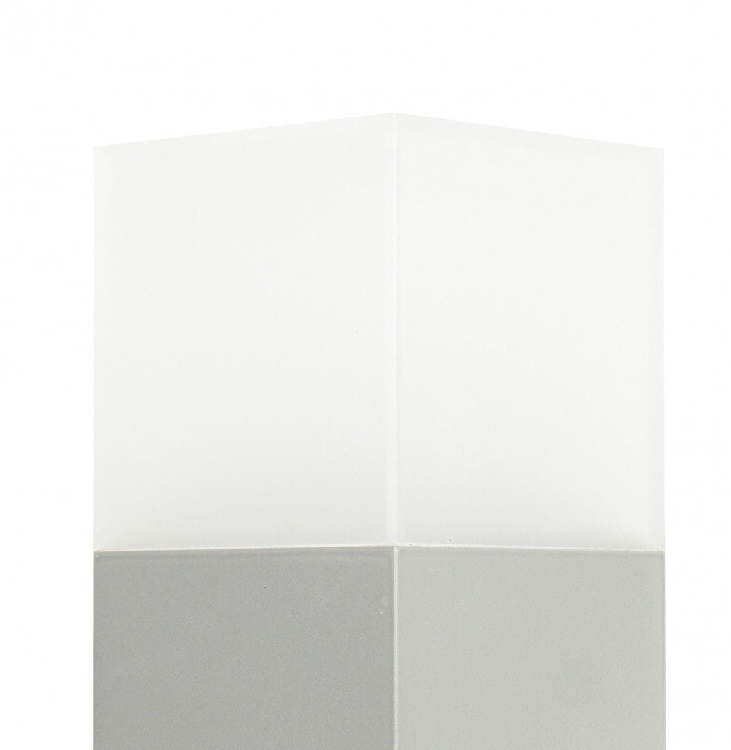 Lampa stojąca ogrodowa Cube, 1-punktowa, wykonana z aluminium. Posiada prostokątną, matową podstawę w kolorze szarym oraz górną część z mlecznego szkła, emitującą światło. Ujęcie z góry, eksponujące kształt i fakturę materiałów.