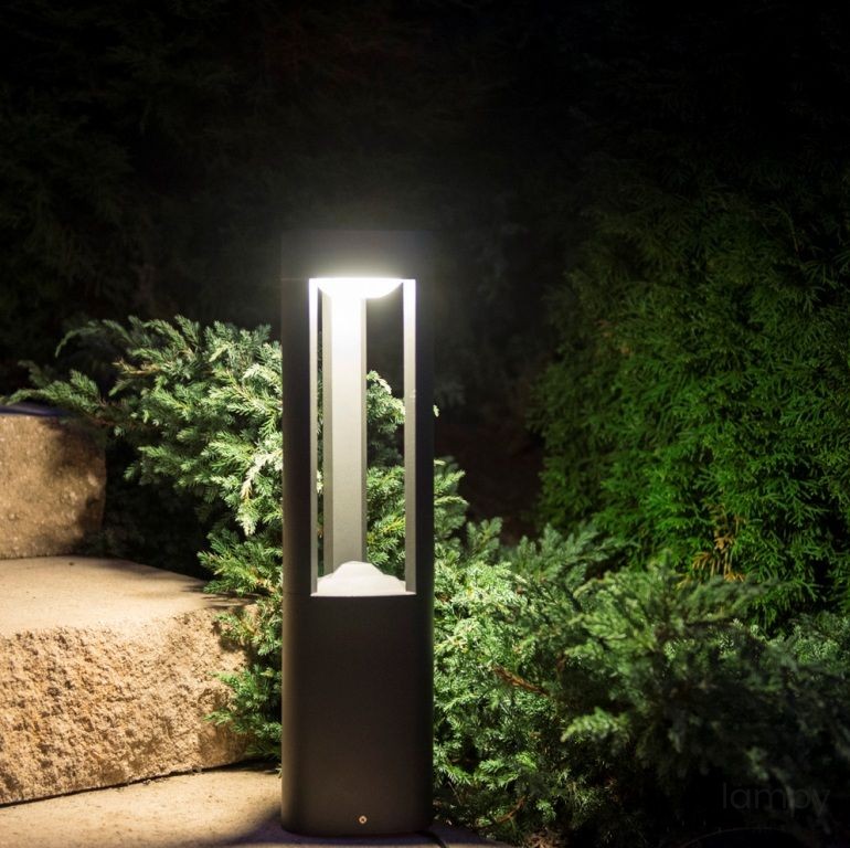 Lampa stojąca ogrodowa 1-punktowa GL11204, czarna, prostokątna, umiejscowiona na kamiennej ścieżce, emitująca ciepłe światło. W tle zieleń krzewów, w nocy. Ustawiony pionowo, z przestrzenią w górnej części, przez którą wydobywa się światło.