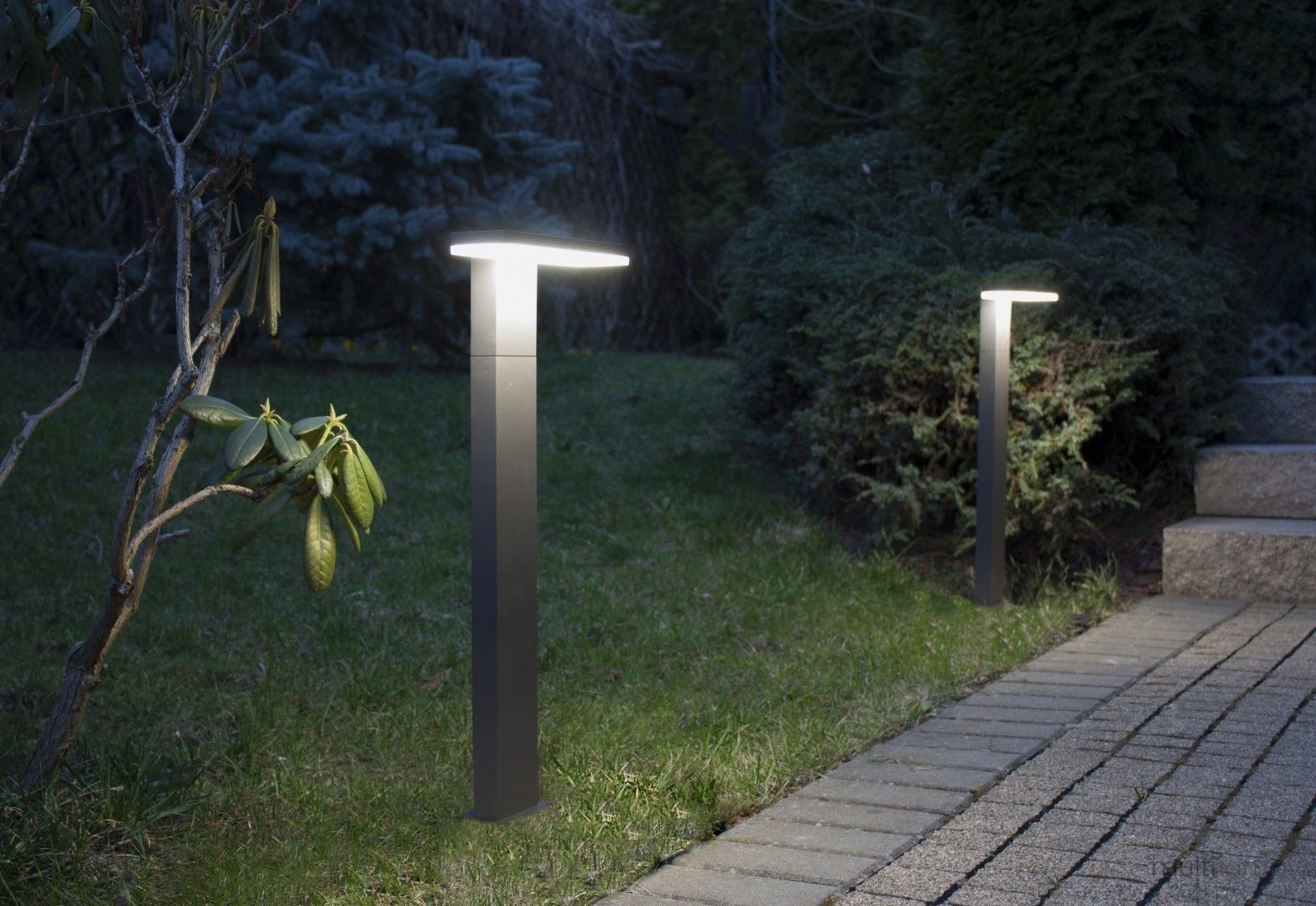 Lampa stojąca ogrodowa Fler H78 LED 10W o barwie 4000K, w kolorze antracytowym, ustawiona na trawie, z prostokątnym kloszem u góry. W tle widoczne krzewy i ścieżka wyłożona kostką brukową. Lampa emituje ciepłe światło, oświetlając otoczenie.