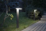 Lampa stojąca ogrodowa Fler H78 LED 10W o barwie 4000K, w kolorze antracytowym, ustawiona na trawie, z prostokątnym kloszem u góry. W tle widoczne krzewy i ścieżka wyłożona kostką brukową. Lampa emituje ciepłe światło, oświetlając otoczenie.