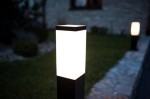 Czarna, kwadratowa lampa stojąca ogrodowa Inox SS802-650 BL, z białym, transparentnym kloszem. Ujęcie z boku, lampa umieszczona na trawniku, w tle widoczny kamienny chodnik i zieleń. Świeci, tworząc delikatne oświetlenie.