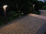 Lampa stojąca ogrodowa Neo H60 z LED 10W, 3000K w ciemnym popiele, umieszczona obok ścieżki. Kąt ujęcia z boku, pokazuje oświetloną powierzchnię chodnika z kostki brukowej w otoczeniu zieleni.