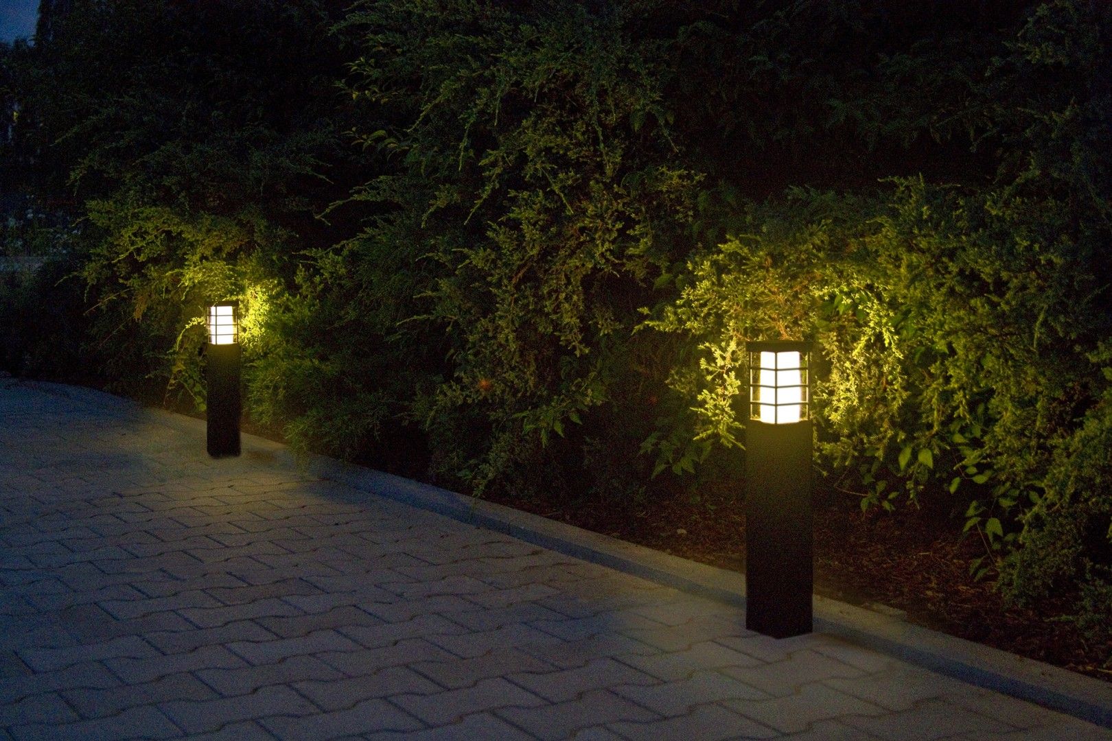 Lampa stojąca ogrodowa Rado II 1-punktowa w kolorze czarnym, o prostokątnym kształcie z białym kloszem z siatką. Ustawiona na podłożu, oświetla ścieżkę w otoczeniu zieleni. Widoczne ciepłe światło wprowadza przytulny nastrój.