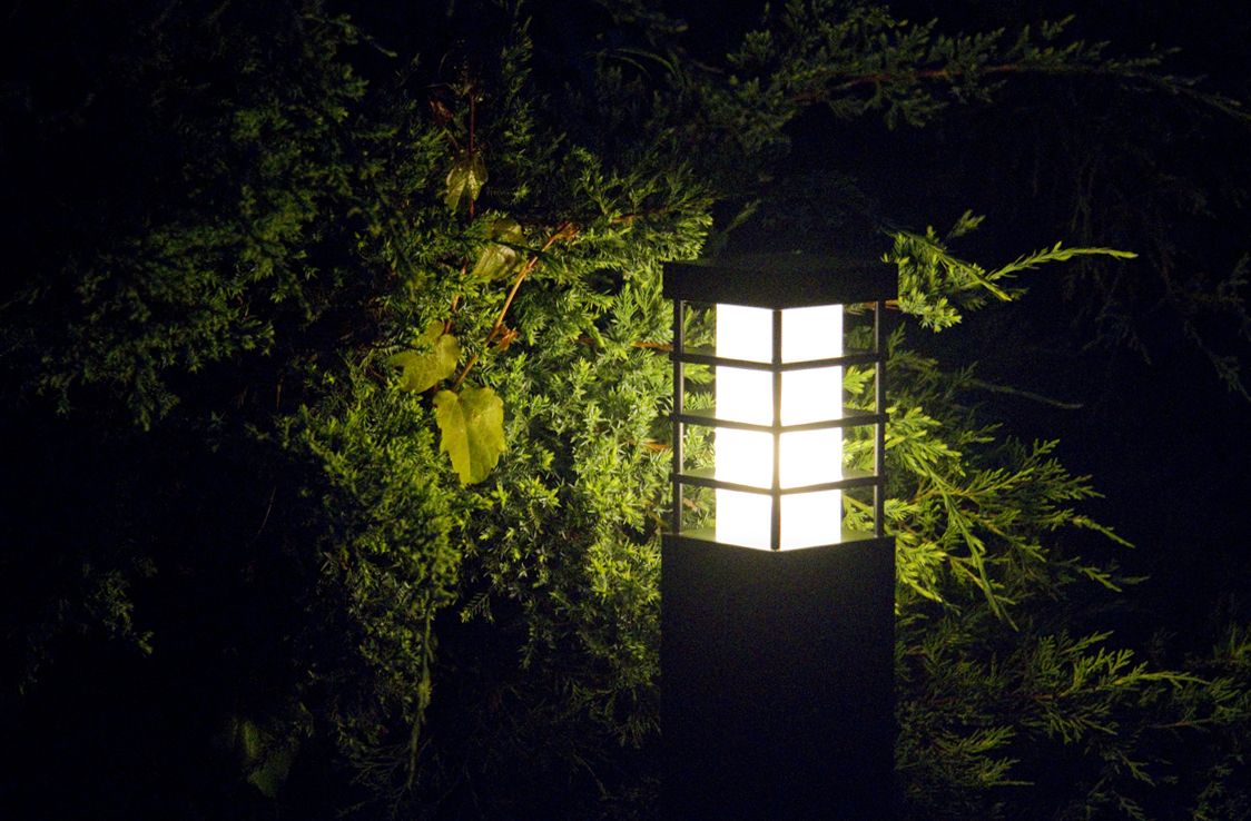 Lampa stojąca ogrodowa Rado II w kolorze czarnym, jednopunktowa, z prostokątnym kloszem z białym szkłem, umieszczona w otoczeniu zieleni. Ujęcie z boku, podkreślające efekt iluminacji w ciemności.