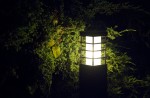 Lampa stojąca ogrodowa Rado II w kolorze czarnym, jednopunktowa, z prostokątnym kloszem z białym szkłem, umieszczona w otoczeniu zieleni. Ujęcie z boku, podkreślające efekt iluminacji w ciemności.