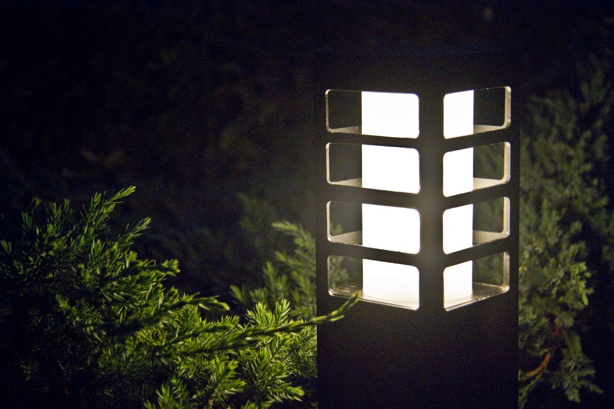 Lampa stojąca ogrodowa Rado III z aluminium, z białym kloszem w formie prostokąta, z widocznymi szczelinami w obudowie. Ujęcie z boku, lampka oświetla otoczenie, umieszczona wśród zieleni, na tle ciemnego otoczenia.