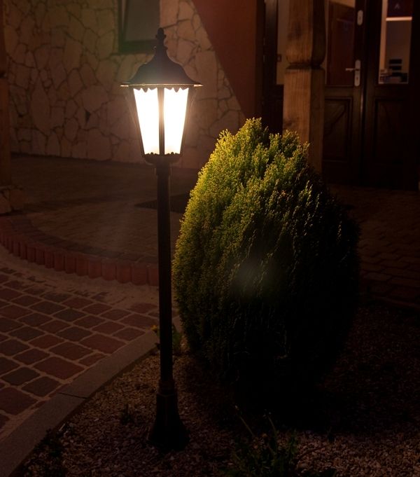 Czarna lampa stojąca ogrodowa Retro Classic H114 z jedną żarówką, o klasycznym designie, umieszczona obok krzewu, w nocy oświetlająca otoczenie. Wysoka podstawa z dekoracyjnym kloszem, na tle kamiennej elewacji budynku.