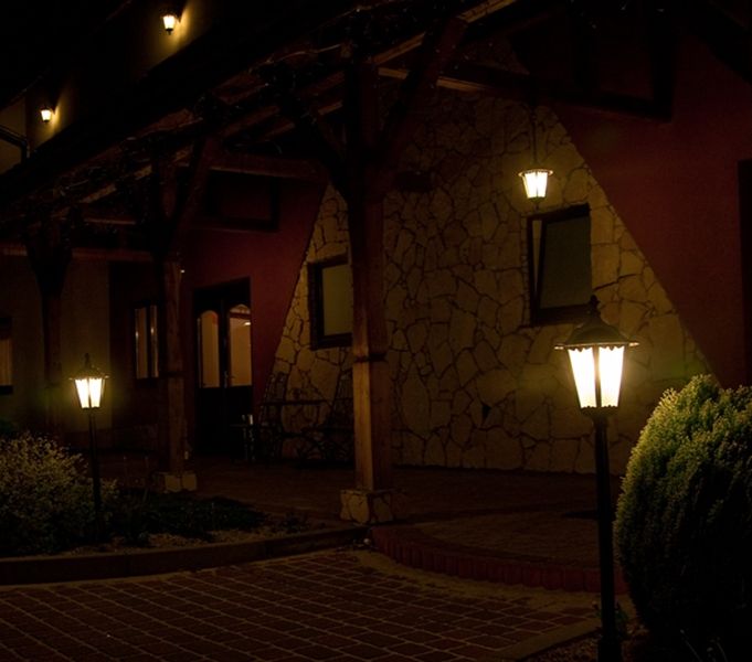 Czarna lampa stojąca ogrodowa Retro Classic H114 z jedną punktową żarówką, umieszczona na tle kamiennej ściany, o klasycznym kształcie z metalowym kloszem. Widoczna w ciemnym otoczeniu, oświetla przestrzeń wokół, tworząc przytulny klimat.