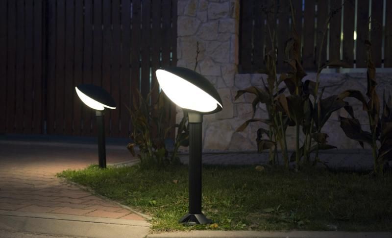 Lampa stojąca ogrodowa Tao 1-punktowa w kolorze czarnym, o nowoczesnym kształcie. Ustawiona na trójnogu na trawniku, emituje jasne światło, z lekko opalizującą, zaokrągloną głowicą, w otoczeniu kamienia i roślinności.