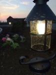 Lampa stojąca ogrodowa Toledo, czarna, 1-punktowa. Szklany klosz z efektownymi kroplami wody, metalowa podstawa z dekoracyjnym ramieniem. Widok z boku, umieszczona w ogrodzie, w tle kwiaty.