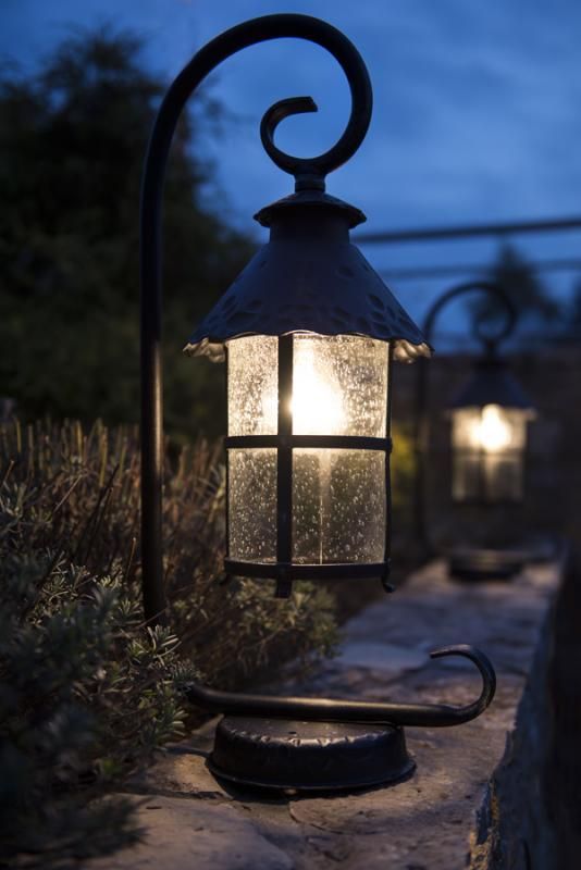 Czarna lampa stojąca ogrodowa Toledo z metalowym stelażem i szklanym kloszem, emitująca ciepłe światło. Ujęcie z boku, lampa ustawiona na kamiennej powierzchni, z dekoracyjnym uchwytem i lekko teksturowanym kloszem. W tle widoczna druga lampa.