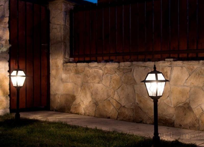 Lampa stojąca ogrodowa Witraż H110, czarna, z jednopunktowym źródłem światła, w kształcie sześciokątnej latarni. Ustawiona na trawniku przy kamiennej ścianie, emituje ciepłe światło w ciemnej scenerii. W tle widoczna drewniana brama.