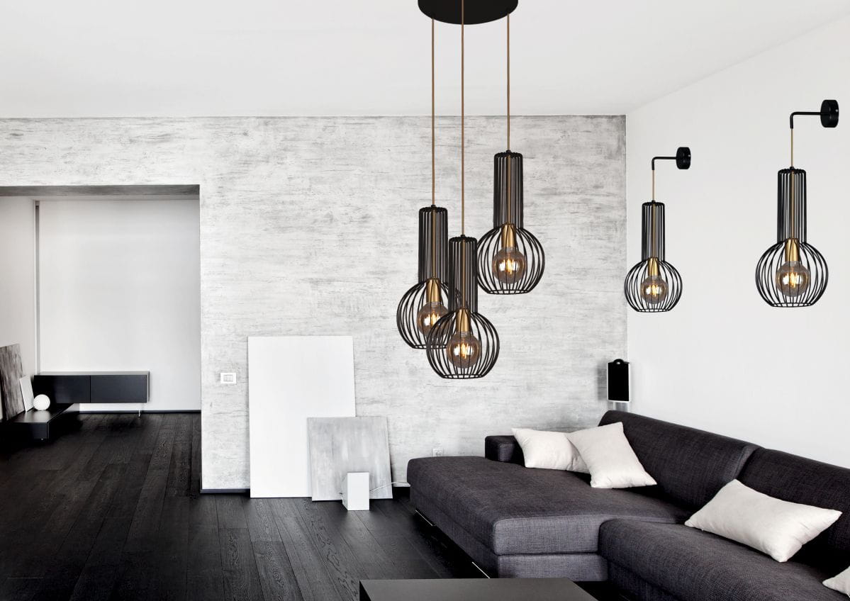 Lampa wisząca Arvi 3-punktowa w kolorze czarnym, z metalowymi kloszami w formie siatki, zawieszona na cienkich linkach. Produkt w nowoczesnym wnętrzu, obok ciemnoszarego sofy i jasnych poduszek. Tło w odcieniach szarości.