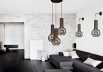 Lampa wisząca Arvi 3-punktowa w kolorze czarnym, z metalowymi kloszami w formie siatki, zawieszona na cienkich linkach. Produkt w nowoczesnym wnętrzu, obok ciemnoszarego sofy i jasnych poduszek. Tło w odcieniach szarości.