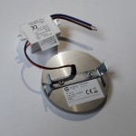 Oti LED oprawa schodowa 230V z ciepłą barwą światła 3000K. Metalowa, okrągła podstawa z widocznym uchwytem montażowym i przewodami. Zasilacz w białej obudowie z oznaczeniami. Produkt przeznaczony do oświetlenia schodów.