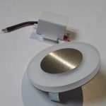 Oti LED oprawa schodowa 230V z okrągłym kloszem z białego tworzywa i metalowym wykończeniem. Widoczny moduł z przewodami. Barwa światła zimna 6500K. Produkt leżący na białym tle, idealny do montażu na schodach lub w korytarzach.