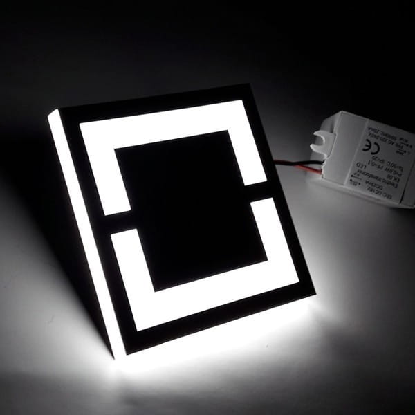 Oprawa schodowa LED 230V o barwie zimnej 6500K, kwadratowa, z czarną ramką i podświetleniem w formie prostokątów. Widoczny zasilacz z przewodami. Ujęcie frontalne, podkreślające minimalistyczny design.