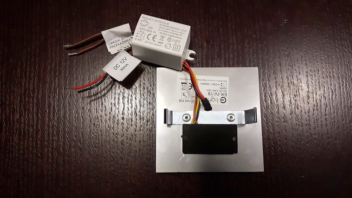 Oprawa schodowa LED z czujnikiem ruchu, 12V, barwa neutralna 4000K. Widoczny moduł sterujący, przewody w kolorach czarnym, czerwonym i brązowym. Elementy metalowe i plastikowe, montażowe mocowanie. Tło ciemne, drewniane.