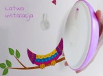 Kinkiet LED Snail w kształcie okrągłym z różową obręczą, trzymany w dłoni, na tle kolorowej naklejki przedstawiającej ptaka na gałęzi. Widoczna informacja łatwa instalacja oraz uchwyt do montażu na ścianie.