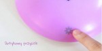 Kinkiet LED Giraffe w formie różowej lampy w kształcie balonu z dotykowym przyciskiem w dolnej części. Na obudowie widoczna ikona słońca. Produkt umieszczony na jasnym tle, z widocznym palcem dotykającym przycisku.