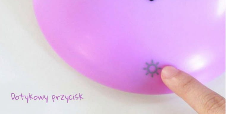 Dotykowy przycisk na różowym, okrągłym kinkiecie Monkey LED, z symbolem słońca, zbliżeniowo wskazywany palcem. W tle widoczny biały stół, co podkreśla minimalistyczny wygląd produktu.