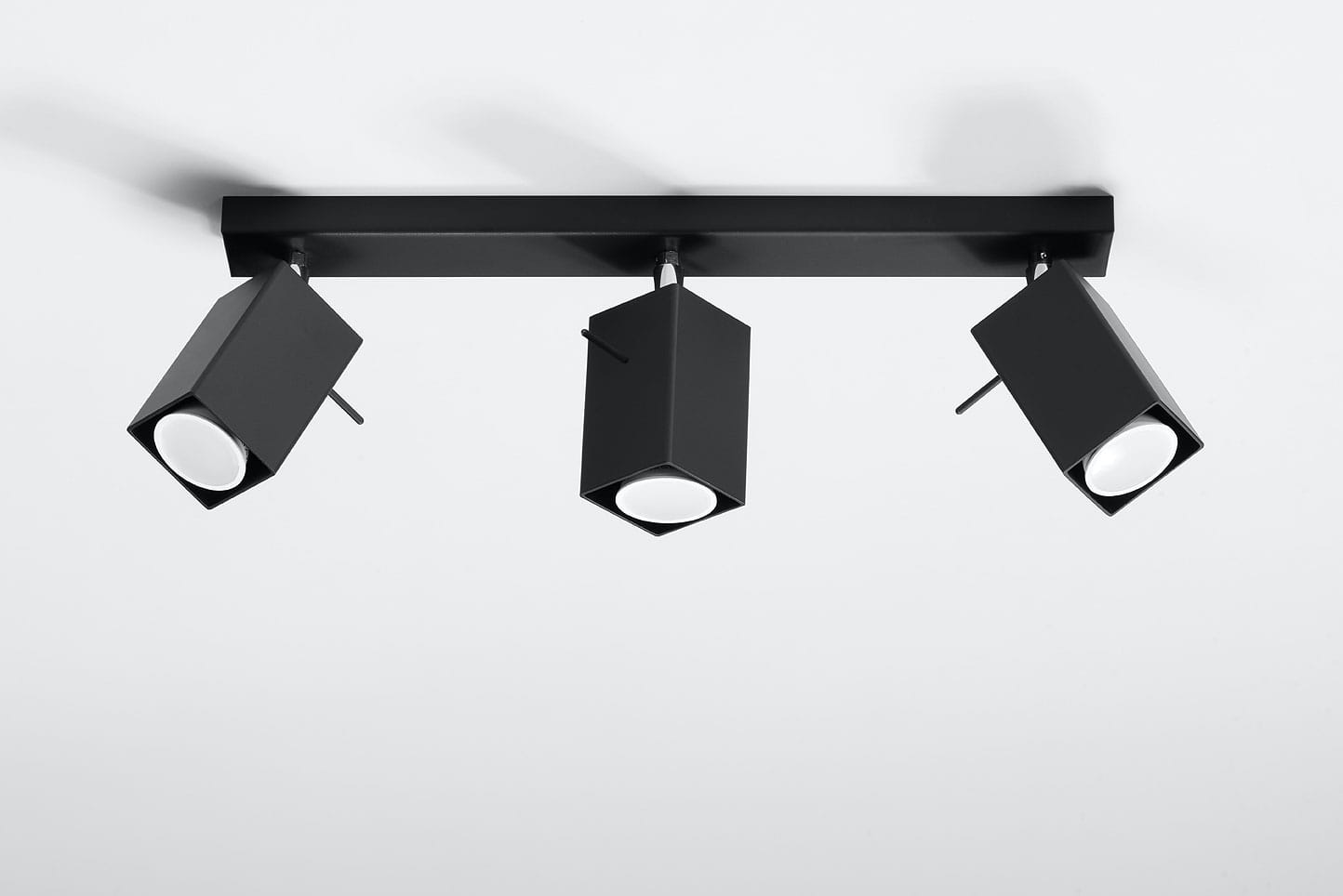 Lampa sufitowa Merida 3-punktowa w kolorze czarnym, z trzema kwadratowymi reflektorami, zamontowana na prostokątnej podstawie. Reflektory są ruchome, co umożliwia kierunkowe oświetlenie. Tło jasne, bez dodatkowych dekoracji.