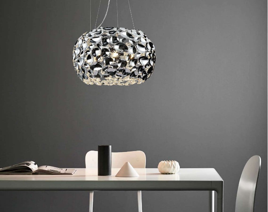 Lampa wisząca Carera Cromo 6-punktowa w chromowanej obudowie, o organicznym kształcie z dekoracyjnymi detalami. Zawieszona nad białym stołem w minimalistycznym wnętrzu, na szarym tle. Widoczne akcesoria: czarny pojemnik, biały wazon, książki.