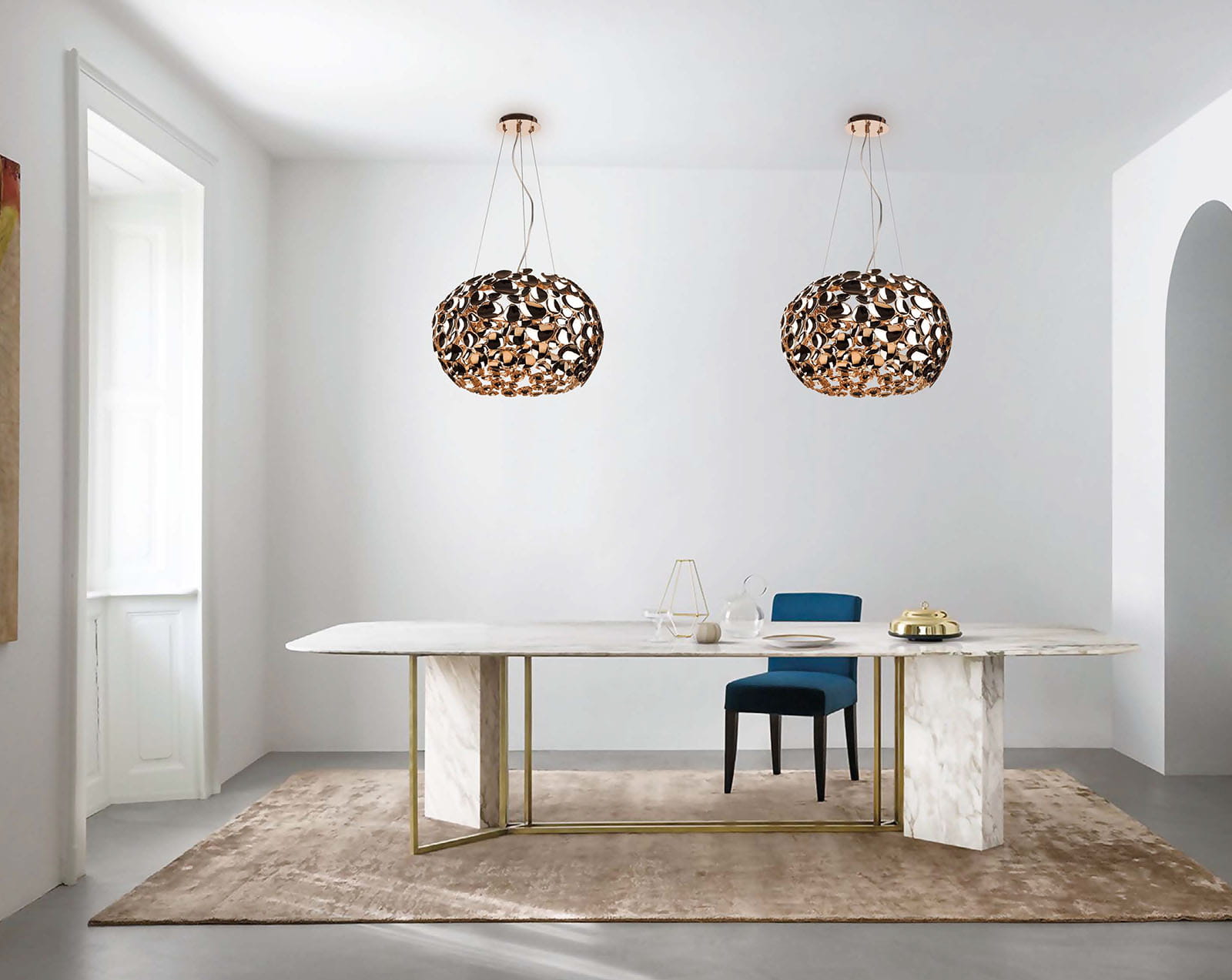 Lampa wisząca Carera Gold z sześcioma punktami świetlnymi w złotym kolorze, o ażurowej strukturze. Zawieszona na długich, cienkich linkach w nowoczesnym wnętrzu z marmurowym stołem i niebieskim krzesłem.