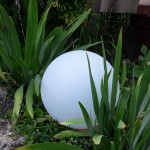 Biała lampa ogrodowa Cumulus M w kształcie kuli, z tworzywa sztucznego, umieszczona na ziemi wśród zielonych roślin. E27, idealna do oświetlenia przestrzeni zewnętrznej. Kąt ujęcia z boku, ukazujący detal i otoczenie.