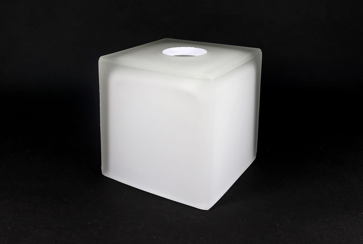 Klosz szklany w kształcie kostki o wymiarach 120x120 mm, wykonany z matowego białego szkła. Posiada okrągły otwór na górze. Produkt stoi na płaskiej powierzchni, na jednolitym ciemnym tle.