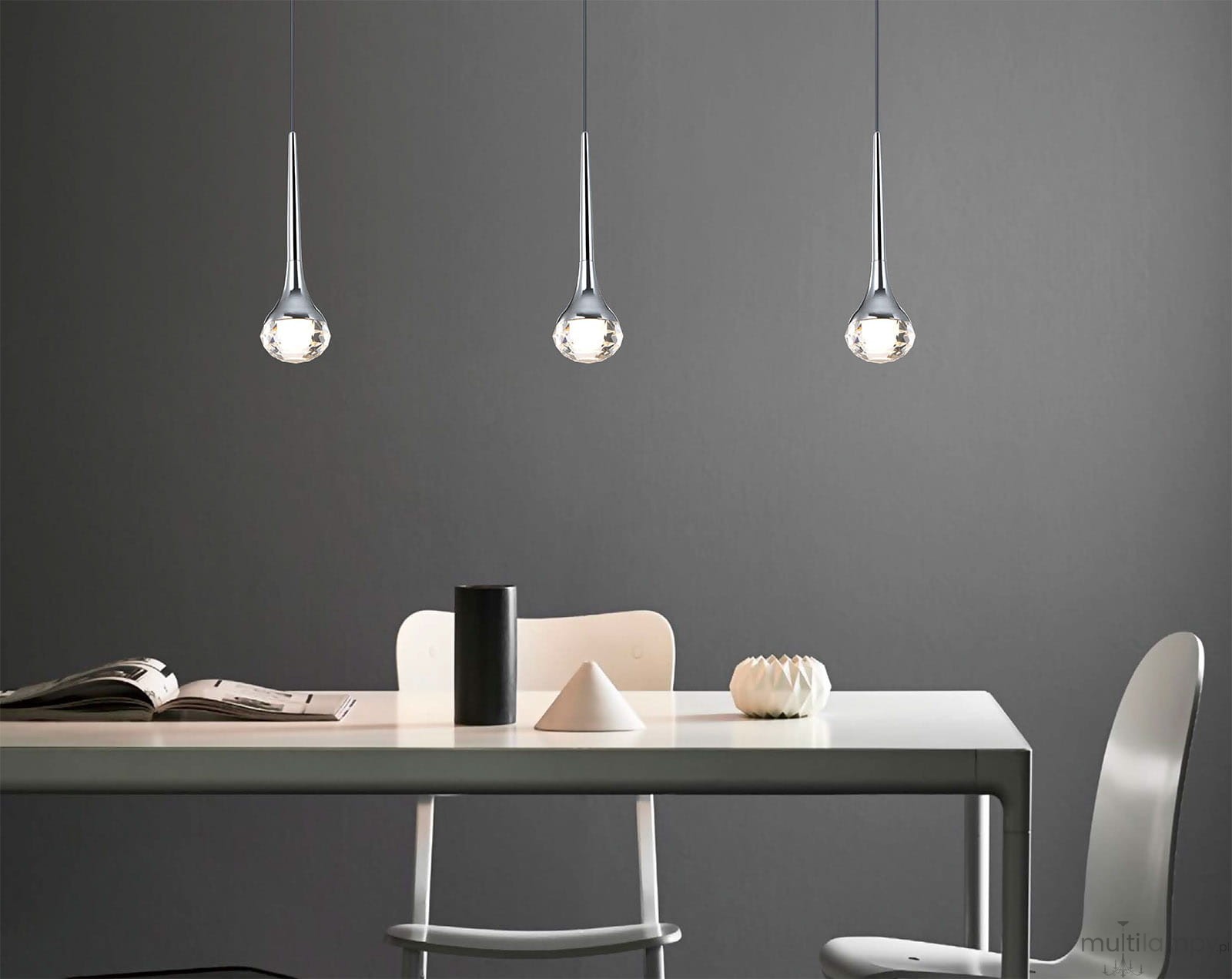 Lampa wisząca Crima Cromo LED w kształcie kropli, metaliczna, z przezroczystym kloszem. Zawieszona nad białym stołem w nowoczesnym wnętrzu, na szarym tle. Widoczne akcesoria na stole: czarny kubek, książka oraz dekoracyjne elementy.