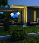 Wisząca lampa ogrodowa Cumulus L, z białym, okrągłym kloszem, zawieszona na cienkim przewodzie. Umieszczona w nowoczesnym otoczeniu, z zielonym trawnikiem i roślinami. W tle widoczny fragment stylowego budynku.