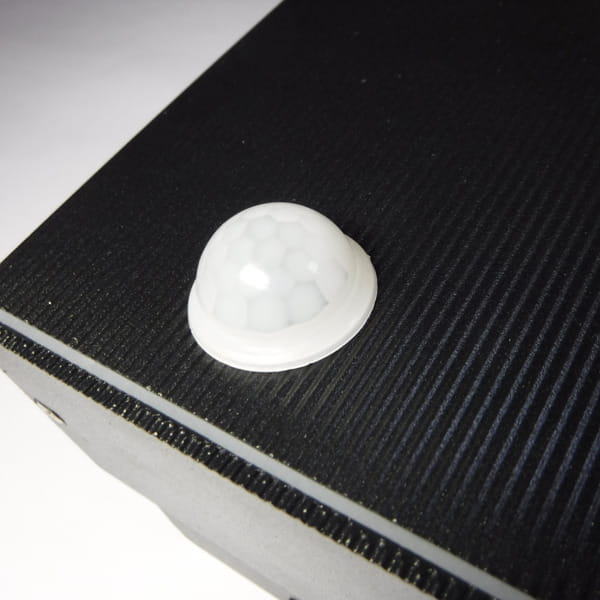 Kinkiet elewacyjny Rio Sensor 1-punktowy z czujnikiem ruchu, zbliżenie na biały czujnik umieszczony na czarnej, teksturowanej powierzchni. Produkt pokazany z góry, z wyraźnym widokiem na strukturę materiału.