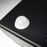 Kinkiet elewacyjny Rio Sensor 1-punktowy z czujnikiem ruchu, zbliżenie na biały czujnik umieszczony na czarnej, teksturowanej powierzchni. Produkt pokazany z góry, z wyraźnym widokiem na strukturę materiału.