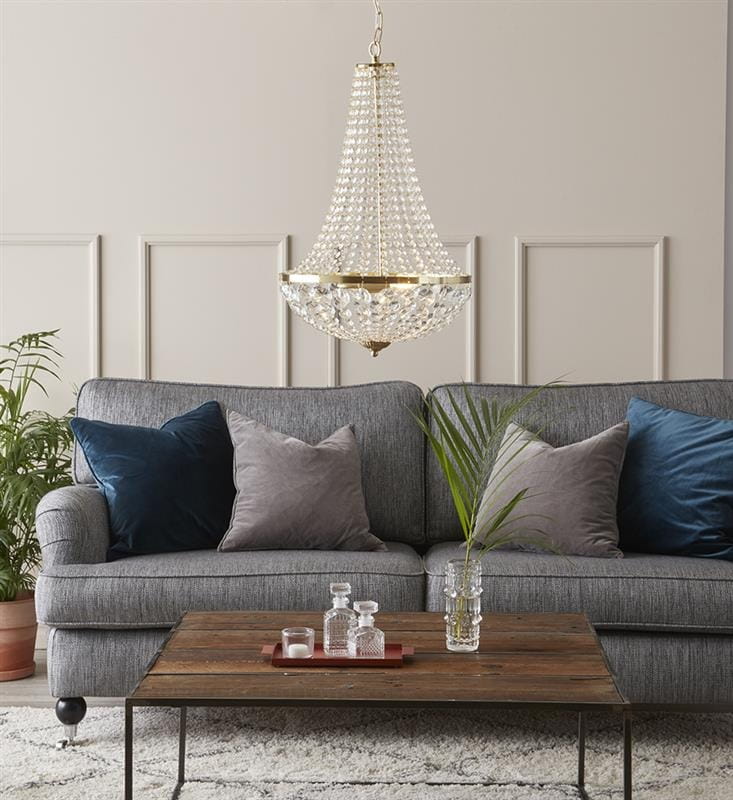 Lampa wisząca Granso 2-punktowa w złotym kolorze, z przezroczystymi kryształkami, zawieszona nad szarym narożnikiem z poduszkami. W tle drewniany stół z szklanymi dodatkami i rośliną, na jasnym tle ściany.