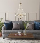 Lampa wisząca Granso 2-punktowa w złotym kolorze, z przezroczystymi kryształkami, zawieszona nad szarym narożnikiem z poduszkami. W tle drewniany stół z szklanymi dodatkami i rośliną, na jasnym tle ściany.