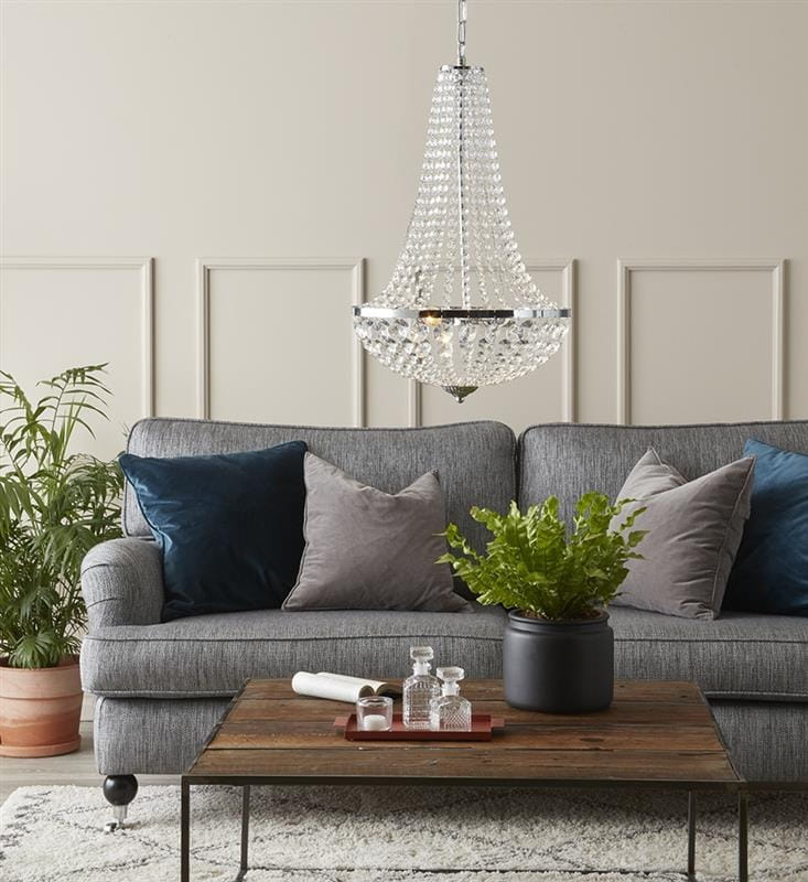 Lampa wisząca Granso 2-punktowa w kolorze chromu, z przezroczystymi kryształami, zawieszona nad szaro-niebieską sofą. W tle widoczne rośliny, drewniany stół z dekoracjami oraz poduszki w różnych odcieniach.