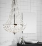 Lampa wisząca Granso 3-punktowa w chromie, z kryształowymi detalami, o kształcie stożkowym. Zawieszona w łazience, na tle białych płytek. Widoczny dwustronny reflektor oraz prostokątne lustro w tle.